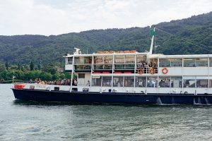 lake-maggiore - Jaya Travel & Tours Passenger ferry sailing on a calm lake with people on the upper deck and forested hills in the background.