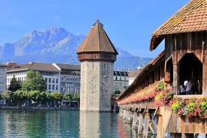 lucerne - Jaya Travel & Tours Lucerne's Chapel Bridge with its stone tower over the Reuss River, flower boxes along the wooden bridge, and the Alps in the background.