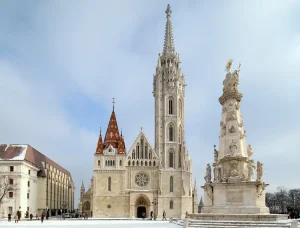 matthias-church - Jaya Travel & Tours Gothic church with a tall central spire and red-tiled towers in a snow-covered square; an ornate monument stands to the right.