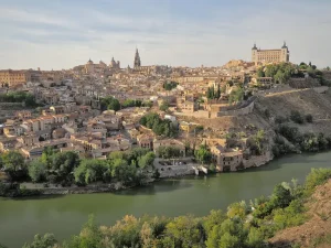medieval-toledo - Jaya Travel & Tours Panoramic view of a historic town along a winding river, with tan stone buildings, a church spire, and a hilltop fortress/ castle in the distance, under a clear sky.