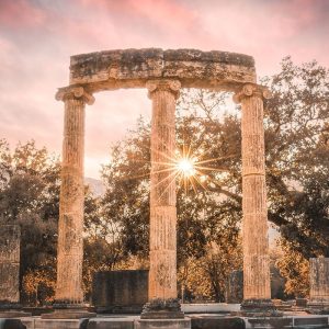 olympia - Jaya Travel & Tours Three ancient stone columns forming a circular ruin at sunset with sun peeking between them.