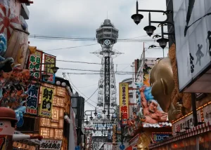 osaka - Jaya Travel & Tours Bustling Osaka street with neon signs and a tall tower in the background; colorful storefronts, banners, and a large golden statue on the right.
