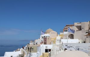 santorini - Jaya Travel & Tours Whitewashed Santorini buildings cascade downslope with blue-domed church, Greek flags fluttering, and the Aegean Sea on the left under a clear blue sky