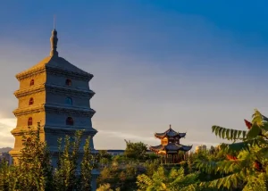 small-wild-goose-pagoda - Jaya Travel & Tours A traditional multi-tier pagoda on the left with a Chinese pavilion in a garden against a sunset sky.