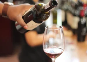 south-african-wine-tour - Jaya Travel & Tours Person pours red wine from a bottle into a stemmed wine glass at a bar or restaurant; blurred background indicates setting.