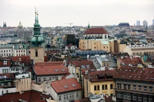 the-city-of-a-thousand-spires - Jaya Travel & Tours Panoramic view of a European city with red-tiled roofs, historic buildings, and green-domed church spires against a pale sky.