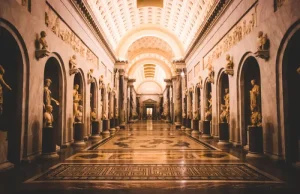 vatican-museums - Jaya Travel & Tours Grand marble gallery with arches and life-size statues along both walls, a polished patterned floor, and a high illuminated coffered ceiling, viewed from a long central aisle.