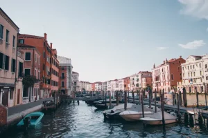 venice - Jaya Travel & Tours Venice canal lined with colorful buildings and docked boats along wooden piers under a clear blue sky, daytime scene.