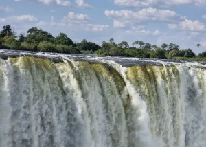 victoria-falls - Jaya Travel & Tours Wide waterfall cascading over a rocky edge with mist, green trees beyond and a blue sky above.
