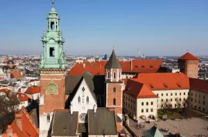 wawel-royal-castle-in-krakow - Jaya Travel & Tours Aerial view of a historic castle complex with red-tiled roofs and a tall green-domed tower against a city skyline.