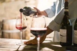 wine-tasting - Jaya Travel & Tours A person pours red wine from a bottle into a glass on a rustic wooden table, with wine bottles in the background.",