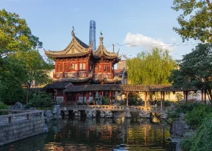yuyuan-garden - Jaya Travel & Tours Traditional Chinese pavilion over a pond with a stone bridge and lush garden, with a modern skyscraper in the background.