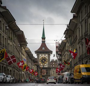 zytglogge-clock-tower - Jaya Travel & Tours View down a cobblestone street toward Bern's ornate clock tower, with flags lining both sides and cars parked along the curb.