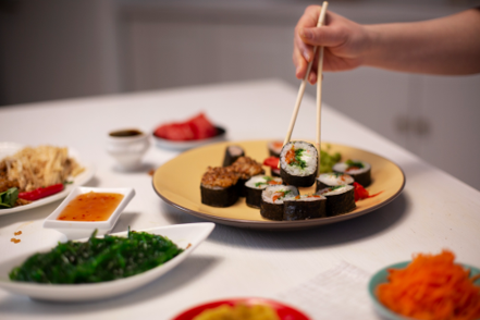 Person uses chopsticks to pick up a sushi roll from a platter of assorted sushi on a table with side dishes and sauces nearby.
