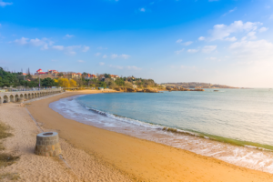 Sunny beach scene with a stone promenade and arches along the coast, and a hillside town in the distance under a blue sky.