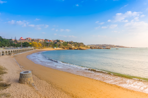 Sunny beach scene with a stone promenade and arches along the coast, and a hillside town in the distance under a blue sky.