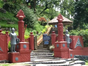 agnigarh-hill-tezpur - Jaya Travel & Tours Red ornate entrance with stone steps leading to a garden, flanked by statues and decorative pillars with carved finials and a thatched pavilion beyond.