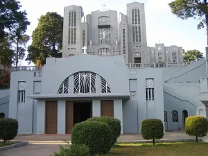 cathedral-of-mary-help-of-christians-shillong-meghalaya - Jaya Travel & Tours Grey art-deco building with twin towers, a central arched window above two wooden doors, and neatly trimmed bushes in front.