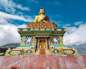 giant-buddha-statue-tawang - Jaya Travel & Tours Golden Buddha statue seated atop a bright, ornate temple pedestal against a blue sky with decorative patterns and dragons on the base.