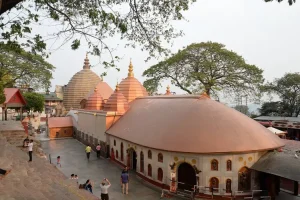 kamakhya-temple-guwahati - Jaya Travel & Tours Temple complex with rounded copper-orange domes and white walls, set in a hillside courtyard as visitors walk along the path under large trees.