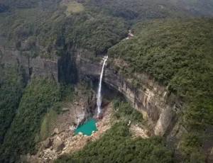nohkalikai-waterfall-cherrapunji - Jaya Travel & Tours A tall waterfall cascades over a cliff into a turquoise pool surrounded by rocky terrain and dense forest.
