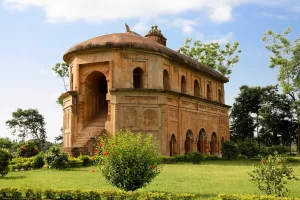 rang-ghar-sivasagar - Jaya Travel & Tours Two-story sandstone pavilion with arches and a curved staircase, set in a manicured garden under a blue sky.