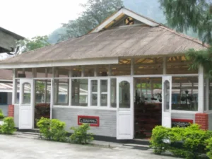 tippi-orchid-centre-arunachal-pradesh - Jaya Travel & Tours Small open-air cafe or shop with white-framed windows and doors, brick base, and green bushes outside.