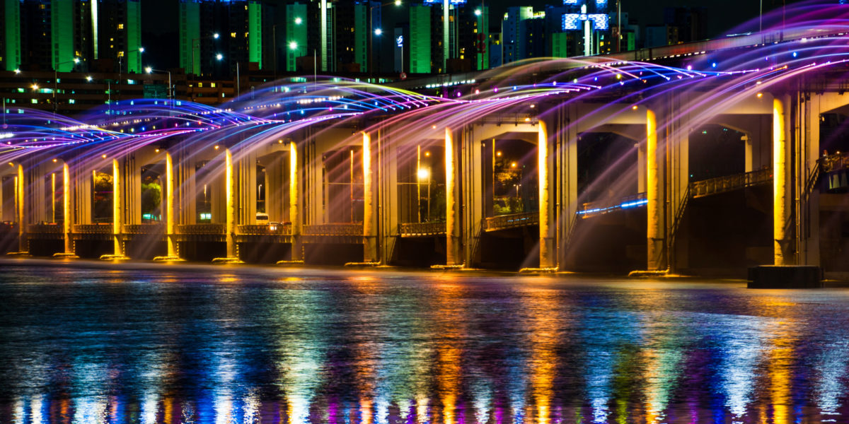 Travel to tour the colored fountain show of the Banpo Bridge Rainbow Fountain against the water reflecting the starlit cityscape of Seoul, South Korea.