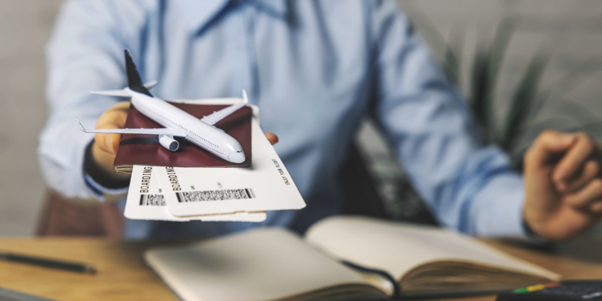 A Jaya Travel & Tours travel agent in a light blue shirt holds a passport and model airplane to demonstrate they just booked a tour for travelers.