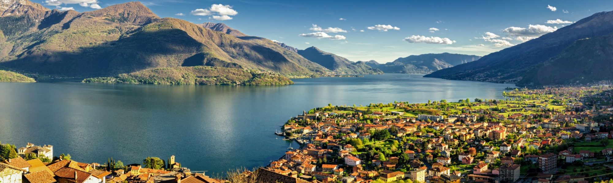 A landscape shot of the beautiful lake como in Gravedona, Italy which is seen on a customized tour from Jaya Travel & Tours. In the image, the sun shines on the small Italian town with the lake and mountains in the background.