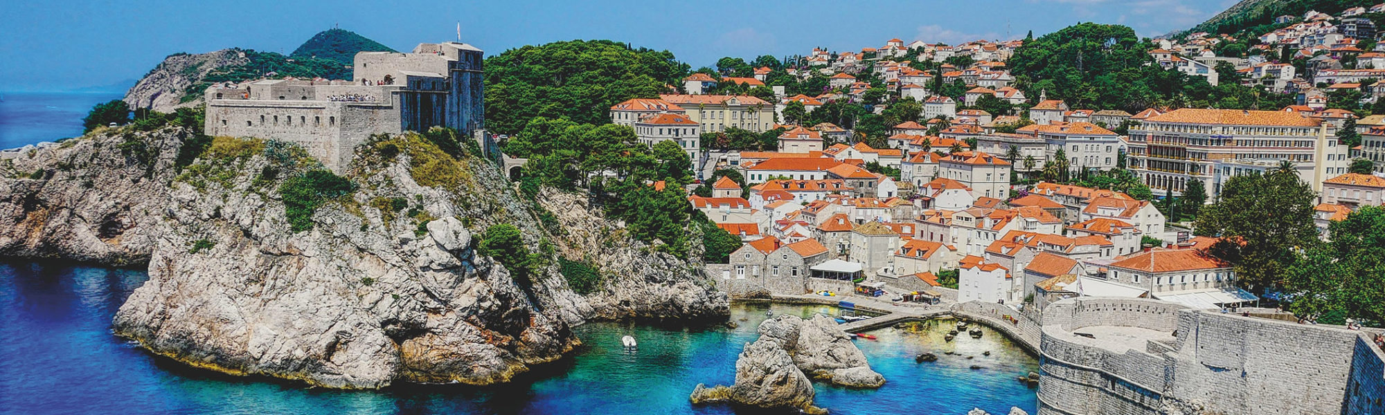 A scenic view of the stone walls of the Walls of Dubrovnik in the west harbor with the ocean and cityscape of Dubrovnik, Croatia from Jaya Travel & Tours.