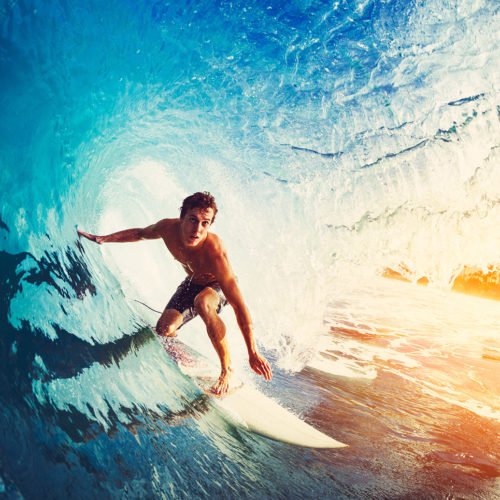 A traveler on a surf board surfs through a blue wave in the ocean on an island hopping adventure with Jaya Travel & Tours.