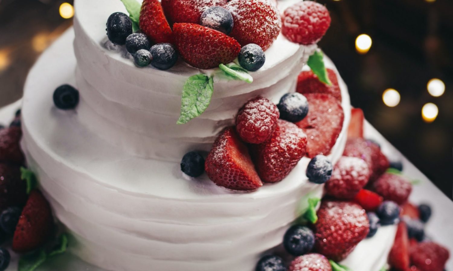 A yummy 3-tier white wedding cake with spatula texture and fresh berries cascading down the side and sprinkled with powdered sugar that you can eat on travel with Jaya.